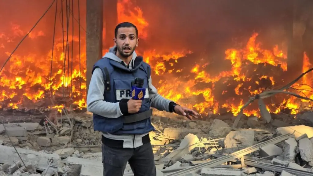 Anas al-Sharif staat met een microfoon van Al Jazeera en een blauw persvest voor een grote brand tussen puin
