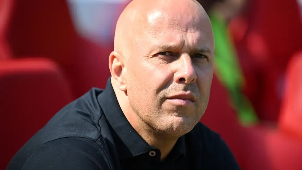 Close-up van trainer Arne Slot in een zwart poloshirt langs de lijn, met rode stadionstoelen op de achtergrond
