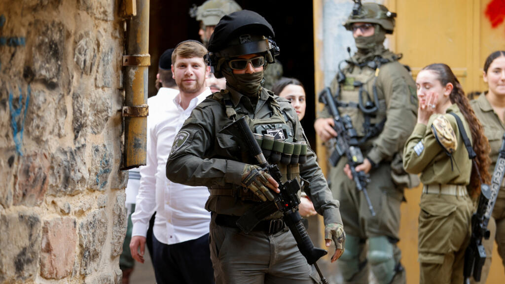 Gewapende Israëlische militairen lopen door een nauwe straat, met kolonisten op de achtergrond