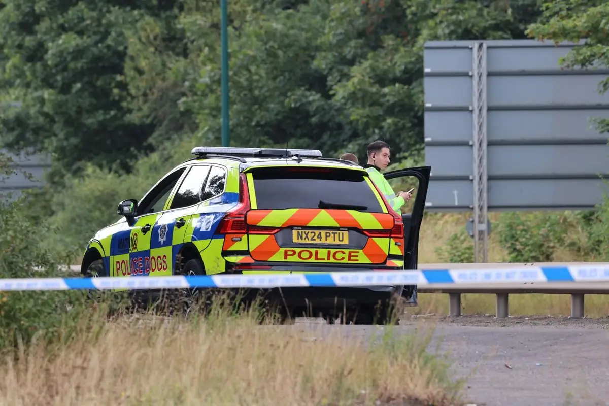 Politieauto met de opdruk police dogs achter blauwwit afzetlint langs de weg, met een agent in geel hesje bij het open portier