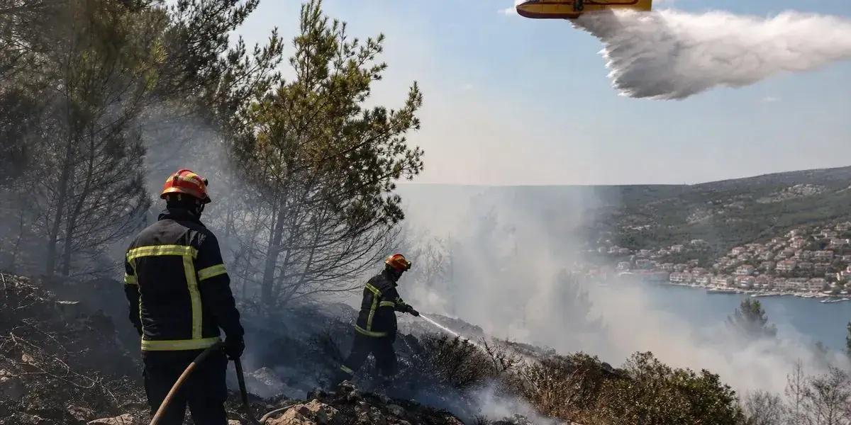 Brandweerlieden blussen tussen rokende bomen terwijl een Canadair-blusvliegtuig water laat vallen, met een kustplaatsje op de achtergrond