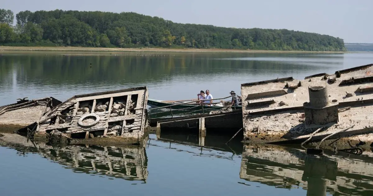 Twee half vergane scheepswrakken uit de Tweede Wereldoorlog steken boven het water uit bij Prahovo, met daarachter mensen in een bootje