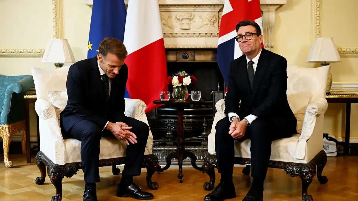 Emmanuel Macron en Andy Burnham zitten naast elkaar in witte fauteuils in Downing Street, met de Europese, Franse en Britse vlag achter zich