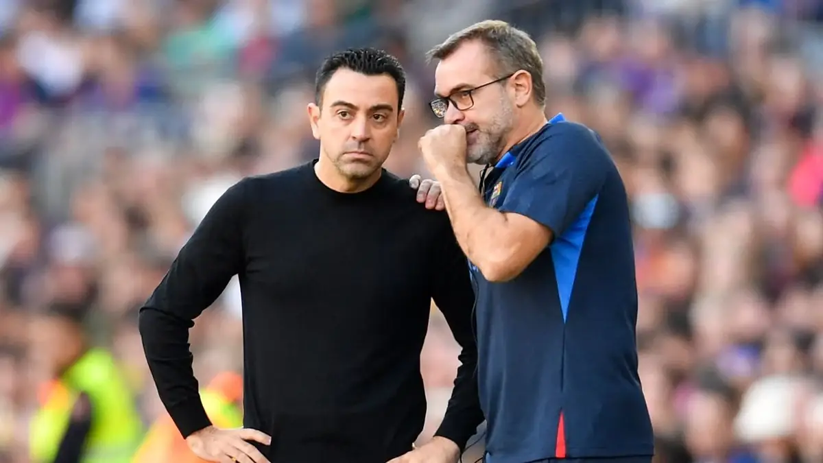 Oscar Hernández praat met zijn hand voor zijn mond in het oor van Xavi langs de lijn, met een vol stadion achter hen