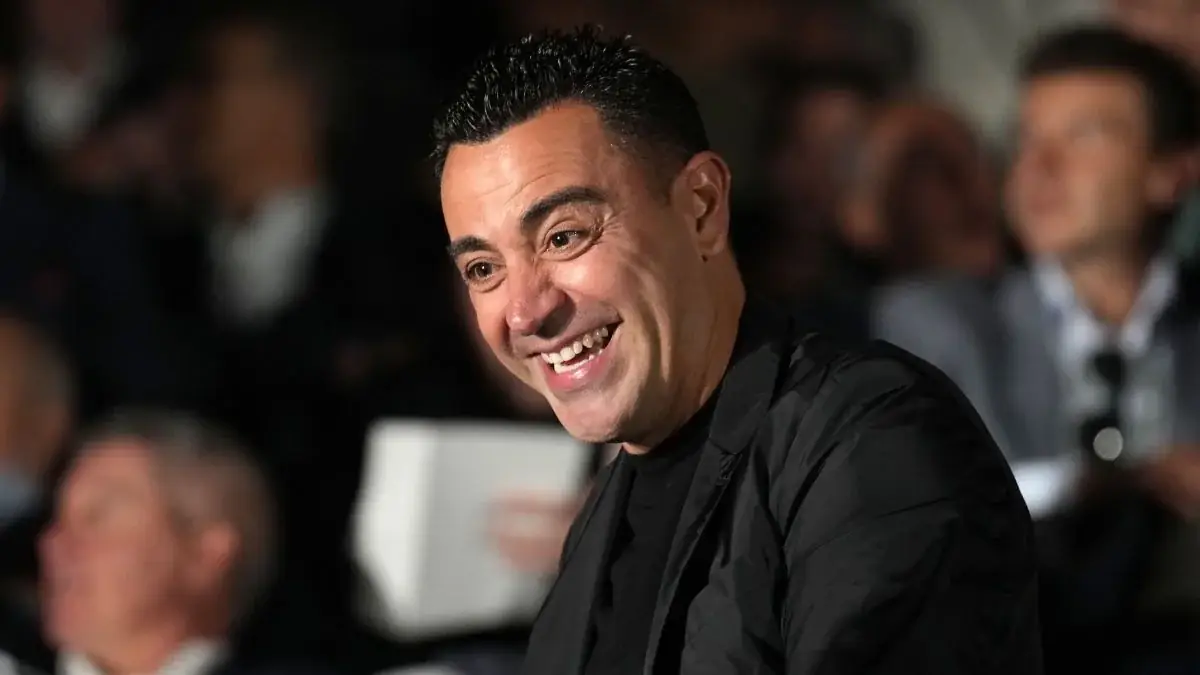 Xavi Hernández lacht in een zwart hemd tussen publiek op een tribune, met wazige gasten op de achtergrond
