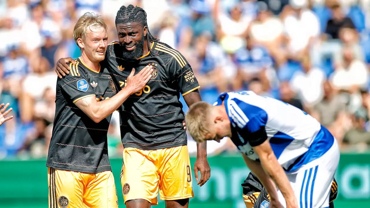 Twee Ajax-spelers in het zwart met gouden uitshirt omhelzen elkaar na een doelpunt, terwijl een tegenstander in een blauw-wit gestreept shirt voorovergebogen staat