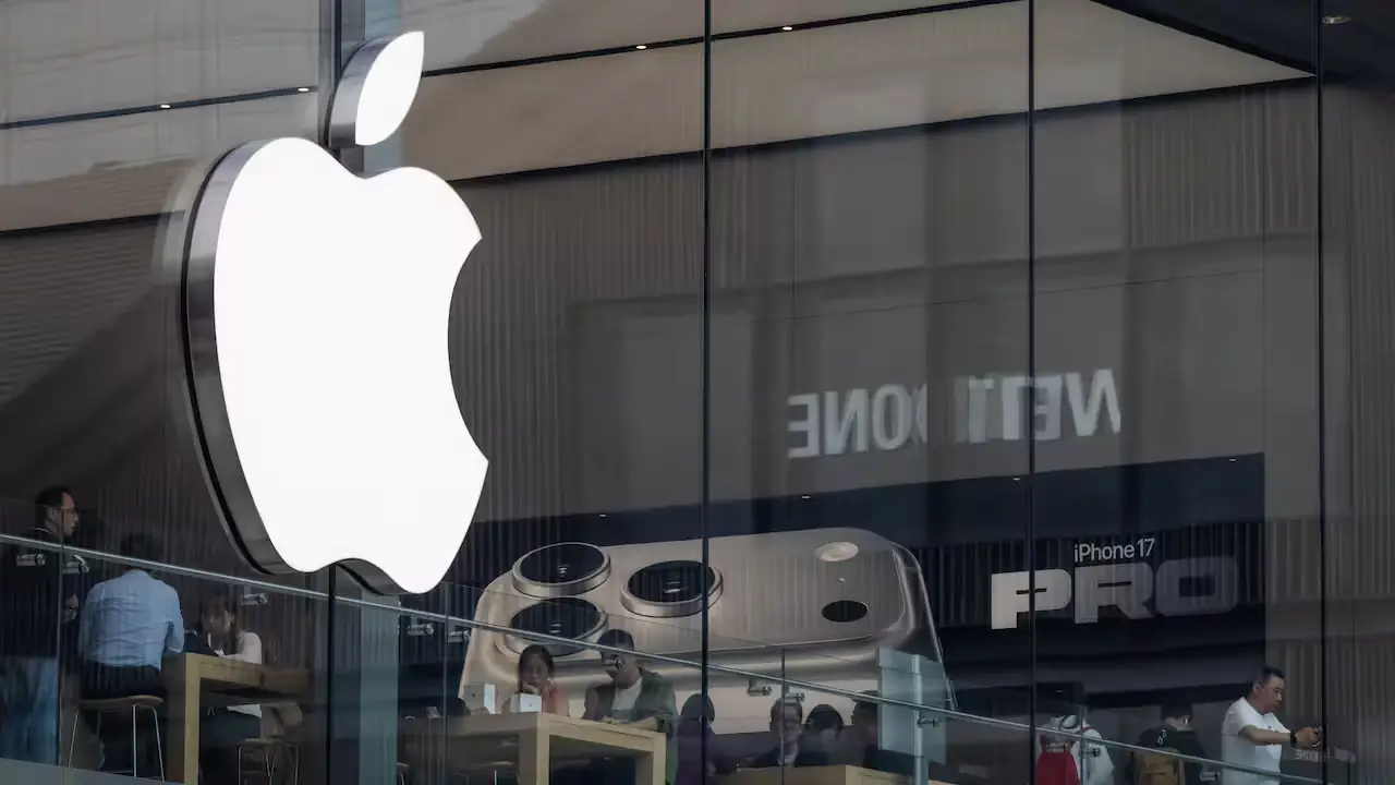 Verlicht Apple-logo aan de glazen gevel van een Apple Store, met daarachter een reclamebeeld voor de iPhone 17 Pro en klanten in de winkel