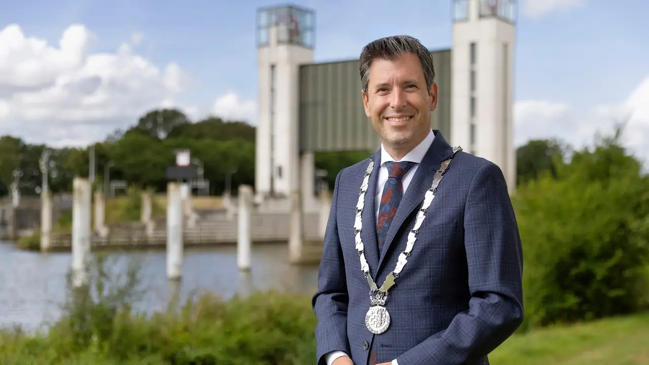 Burgemeester Minses van Heumen in pak met ambtsketen, aan het water met een stuw op de achtergrond