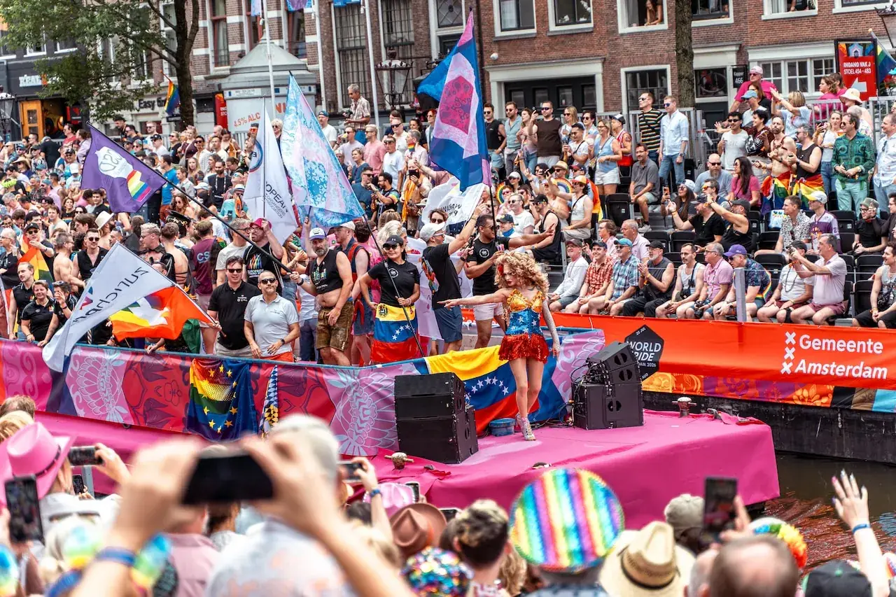 Een boot met regenboogvlaggen en een artiest op het podium vaart door een Amsterdamse gracht, met publiek op de kade en een tribune met de tekst WorldPride Amsterdam 2026