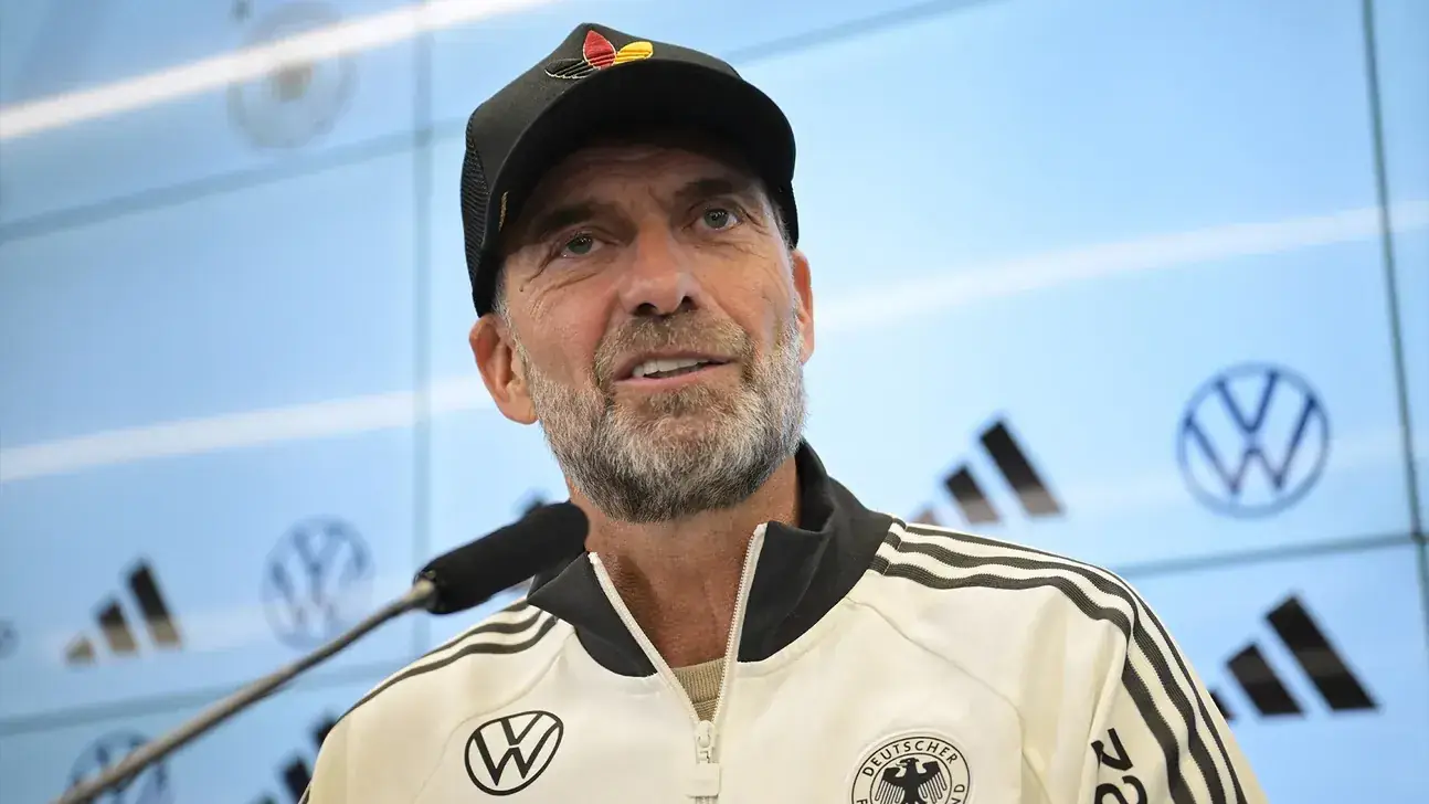 Jurgen Klopp in Duits trainingspak en pet tijdens een persconferentie voor een blauw scherm met sponsorlogos