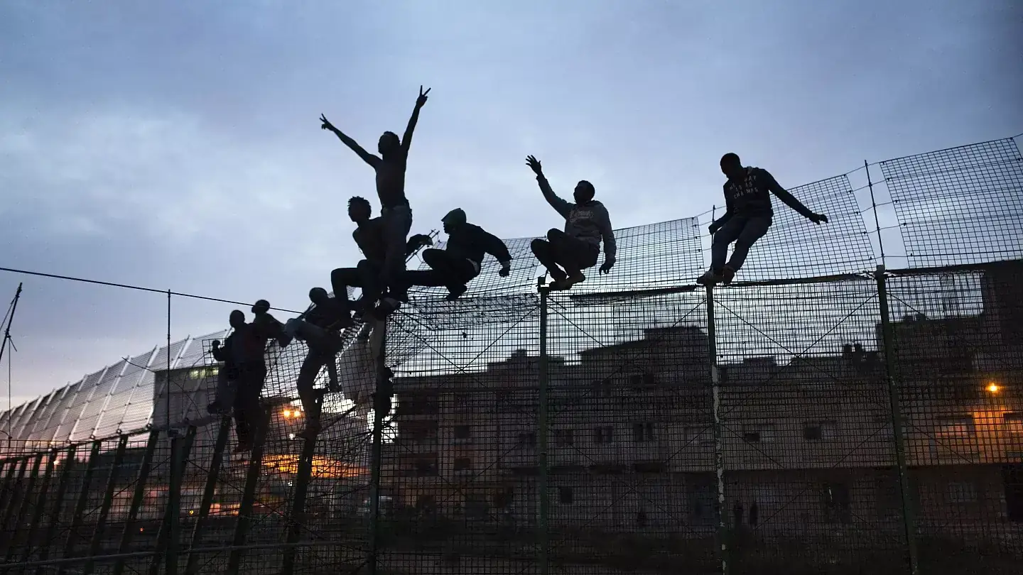 Silhouetten van mensen die in de schemering bovenop het hoge grenshek bij de Spaanse enclave klimmen, met flatgebouwen op de achtergrond