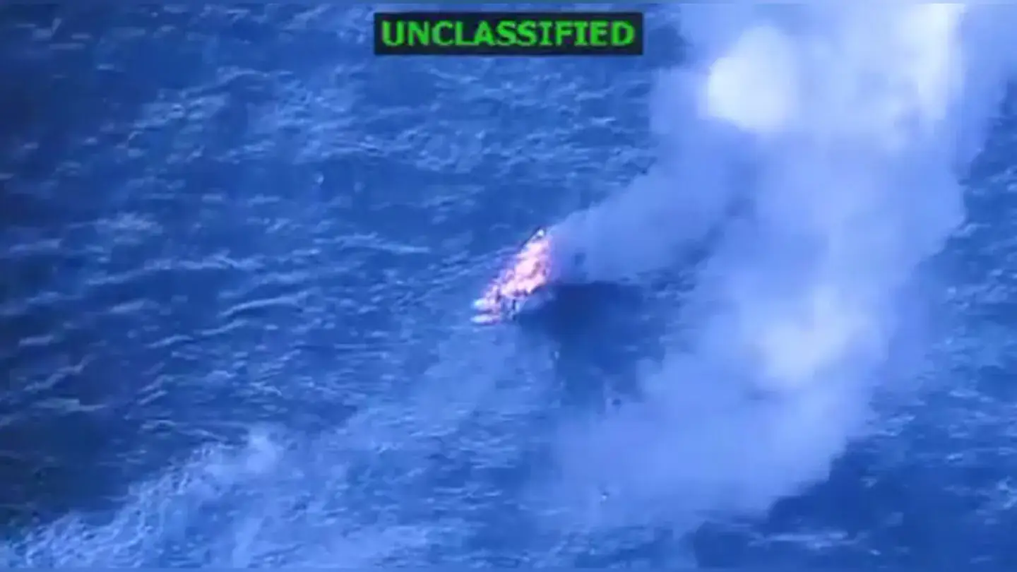 Luchtbeeld van een kleine brandende boot op open zee met een rookpluim, met in de hoek het opschrift unclassified
