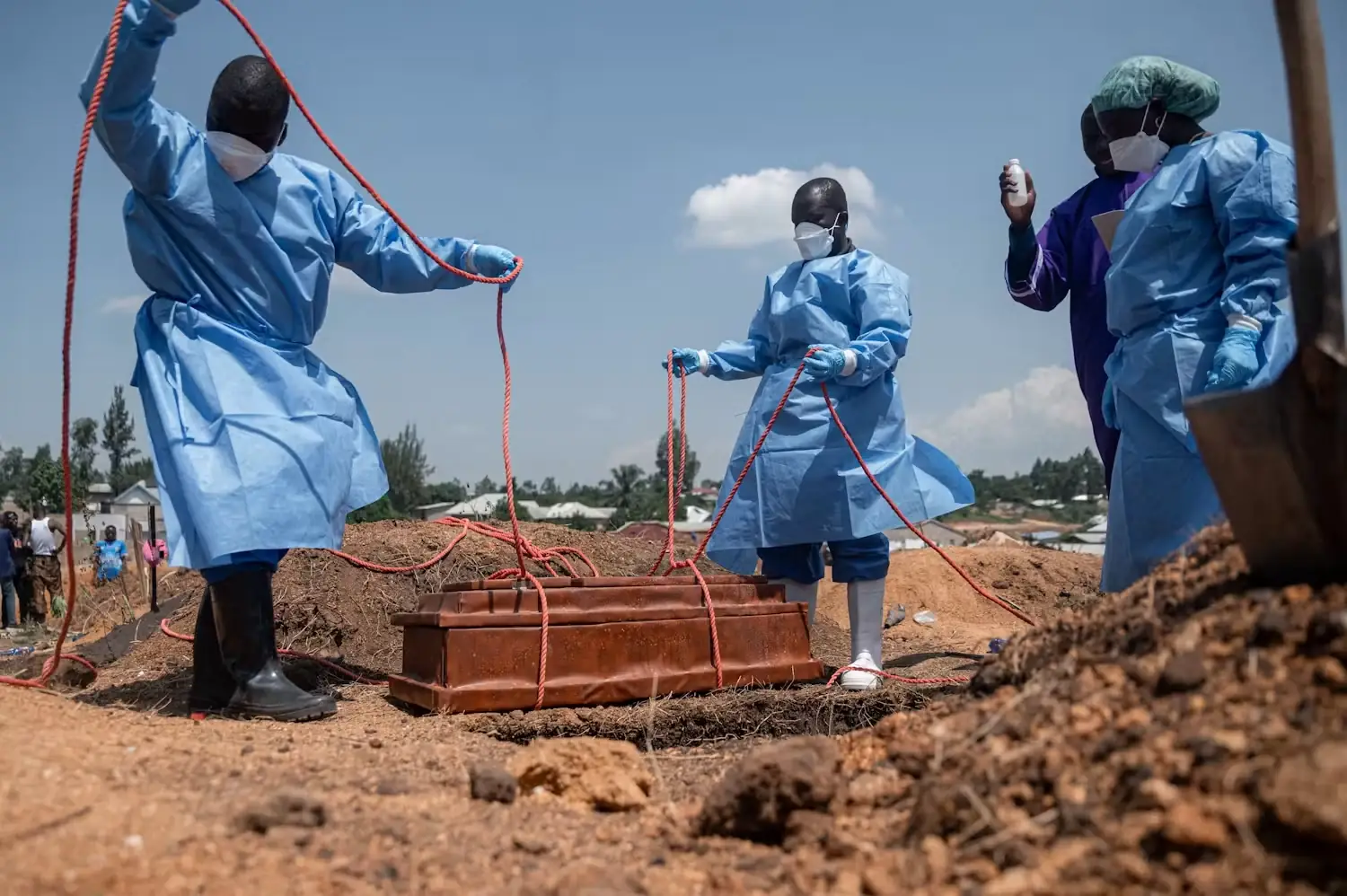 Begrafenismedewerkers in blauwe beschermende pakken laten met touwen een kist in een graf zakken op een begraafplaats in Congo