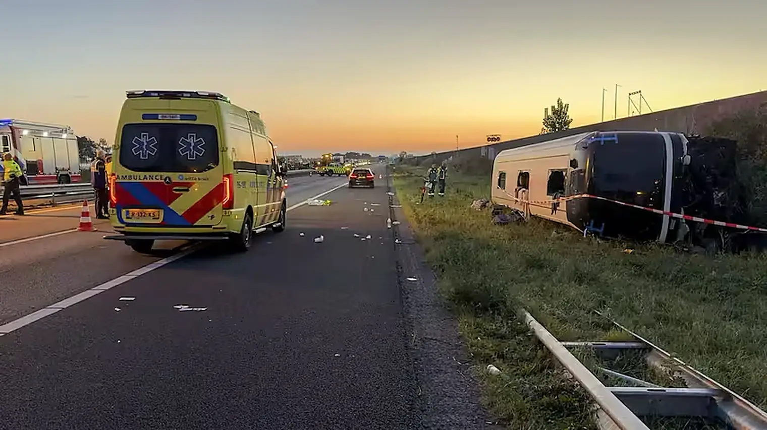 Op zijn kant liggende touringcar in de berm naast de A28, met een ambulance en brokstukken op het asfalt bij zonsopkomst