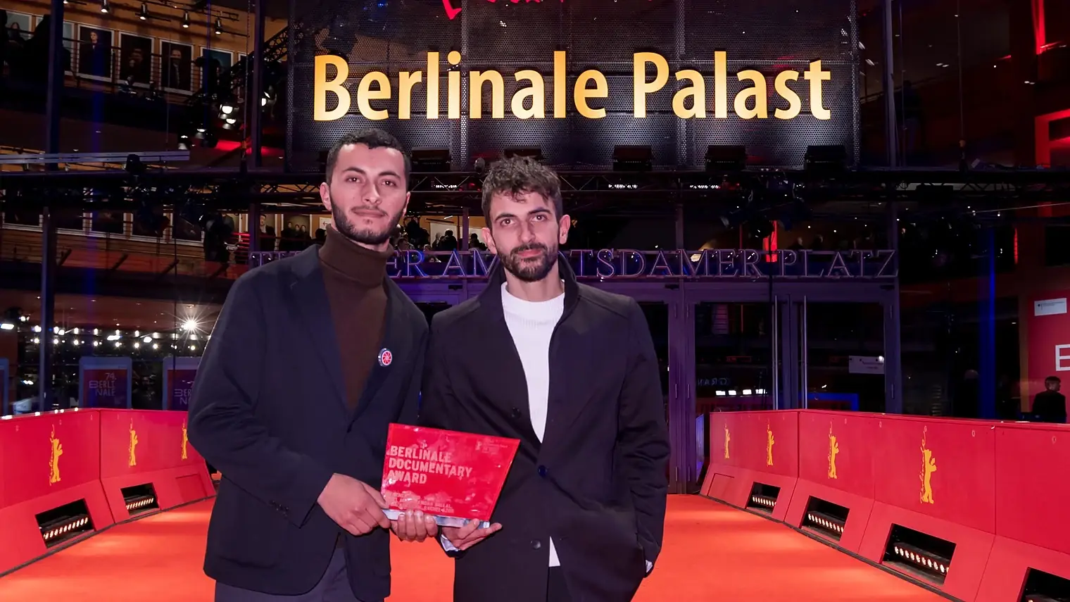 Twee mannen in donkere jassen op de rode loper voor het Berlinale Palast, met tussen hen in een rode prijsoorkonde