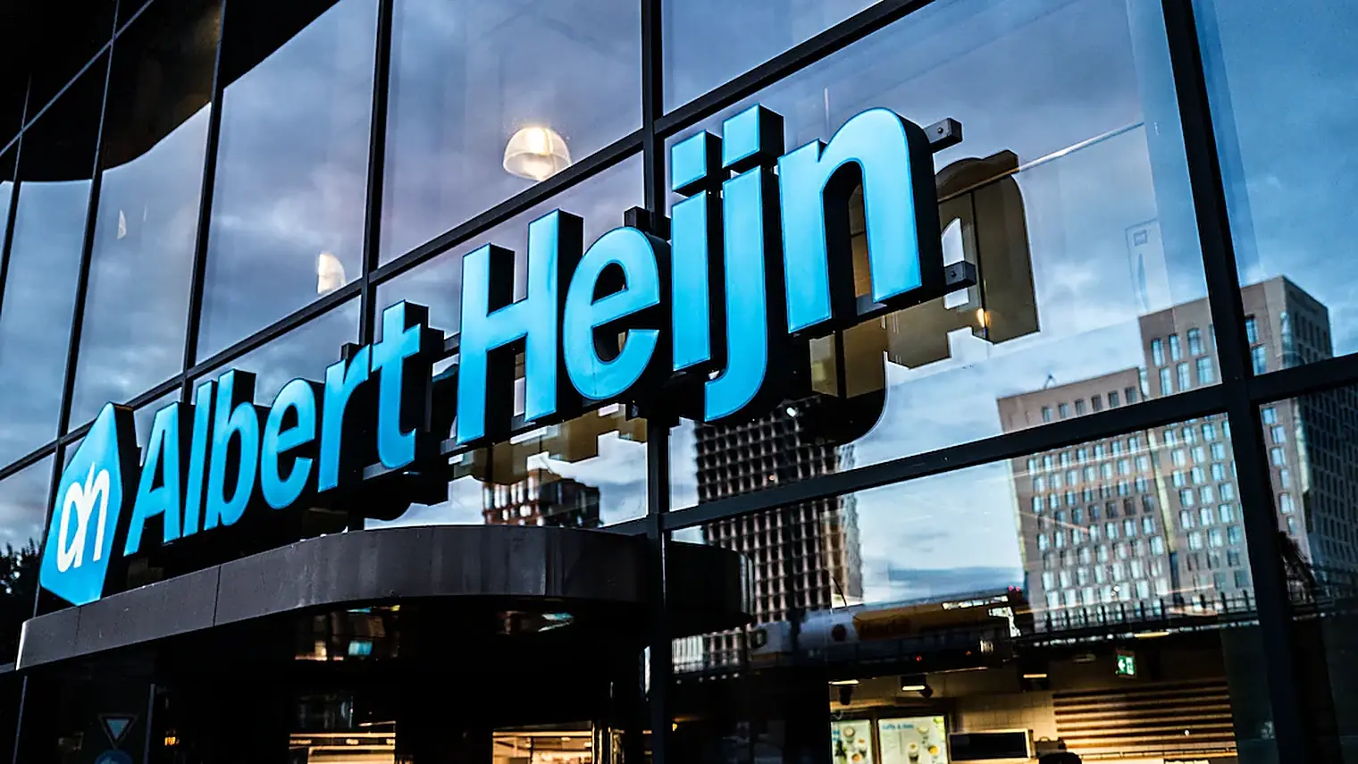 Verlicht Albert Heijn-logo op een glazen gevel, met flatgebouwen weerspiegeld in de ramen