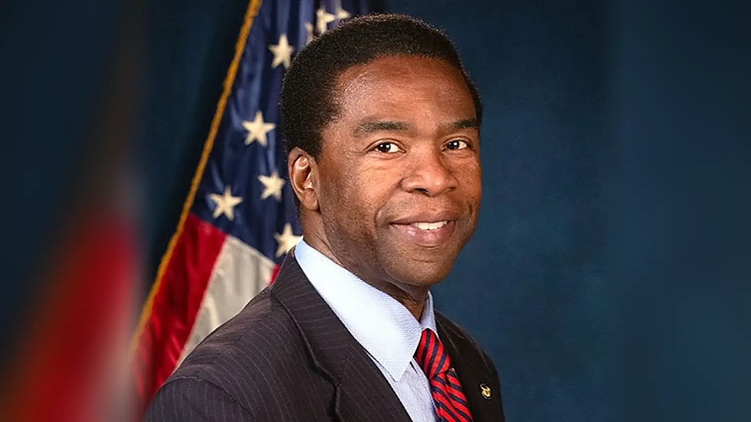 Alvin Brown in pak met gestreepte stropdas voor een Amerikaanse vlag, glimlachend naar de camera