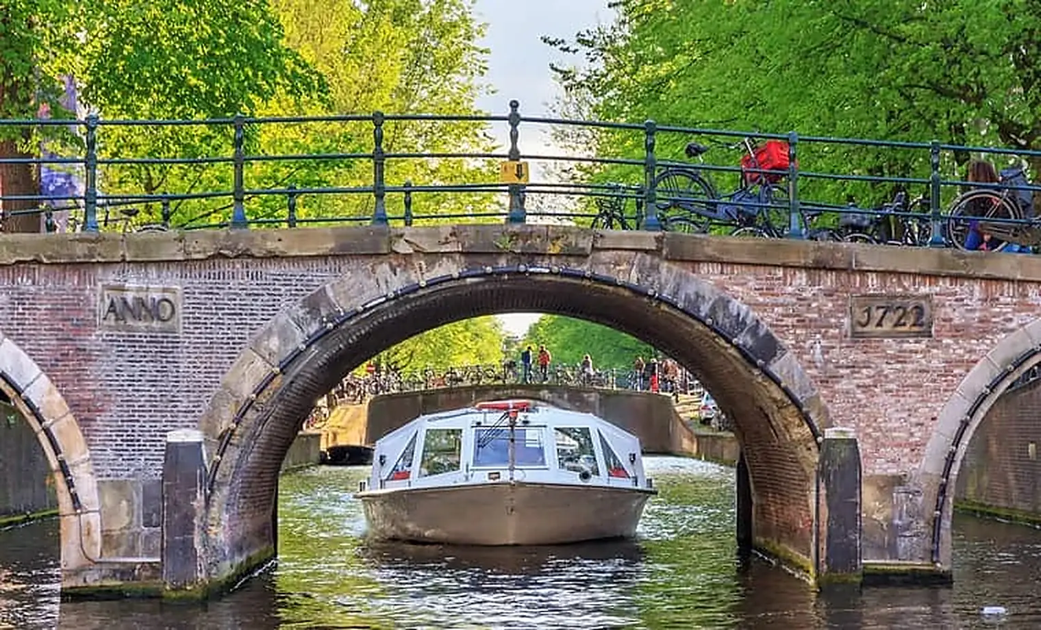 Rondvaartboot met toeristen vaart onder een oude bakstenen boogbrug in een Amsterdamse gracht, met fietsen op de brugleuning