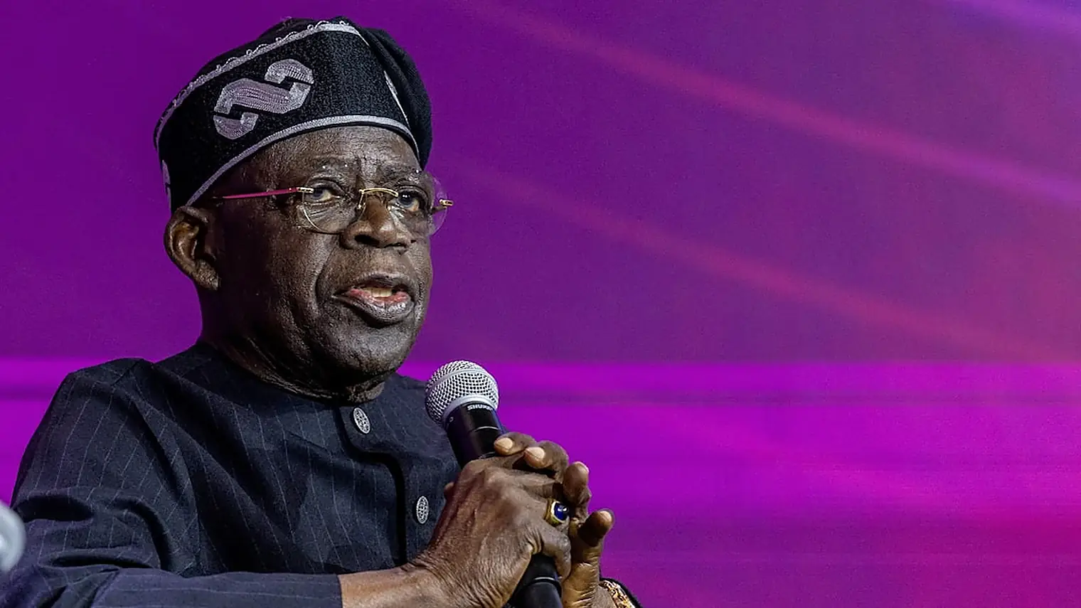 President Bola Tinubu spreekt met een microfoon in de hand, met een paarse achtergrond