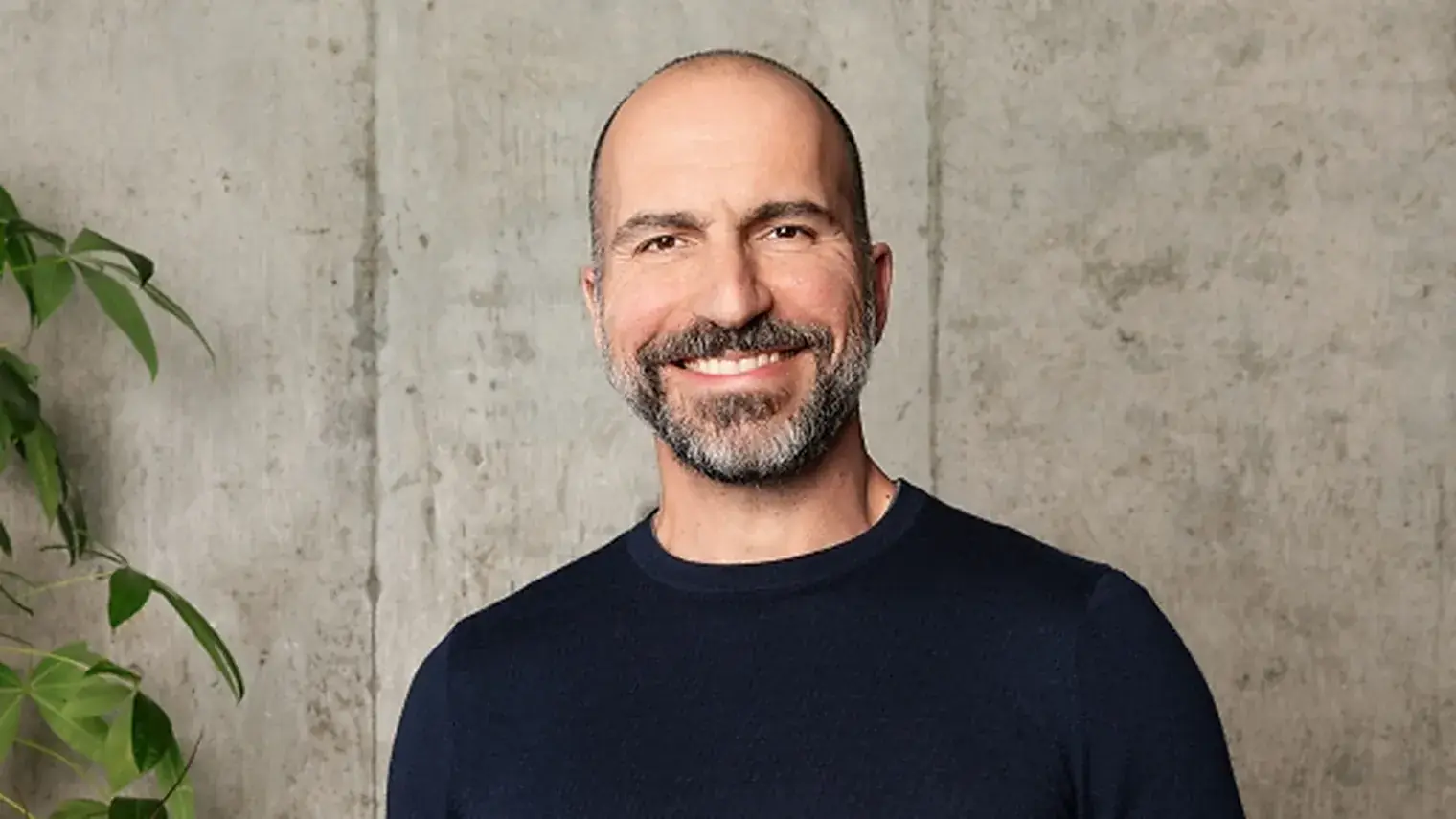 Uber-topman Dara Khosrowshahi in een donkerblauwe trui, lachend voor een betonnen wand met een plant naast hem