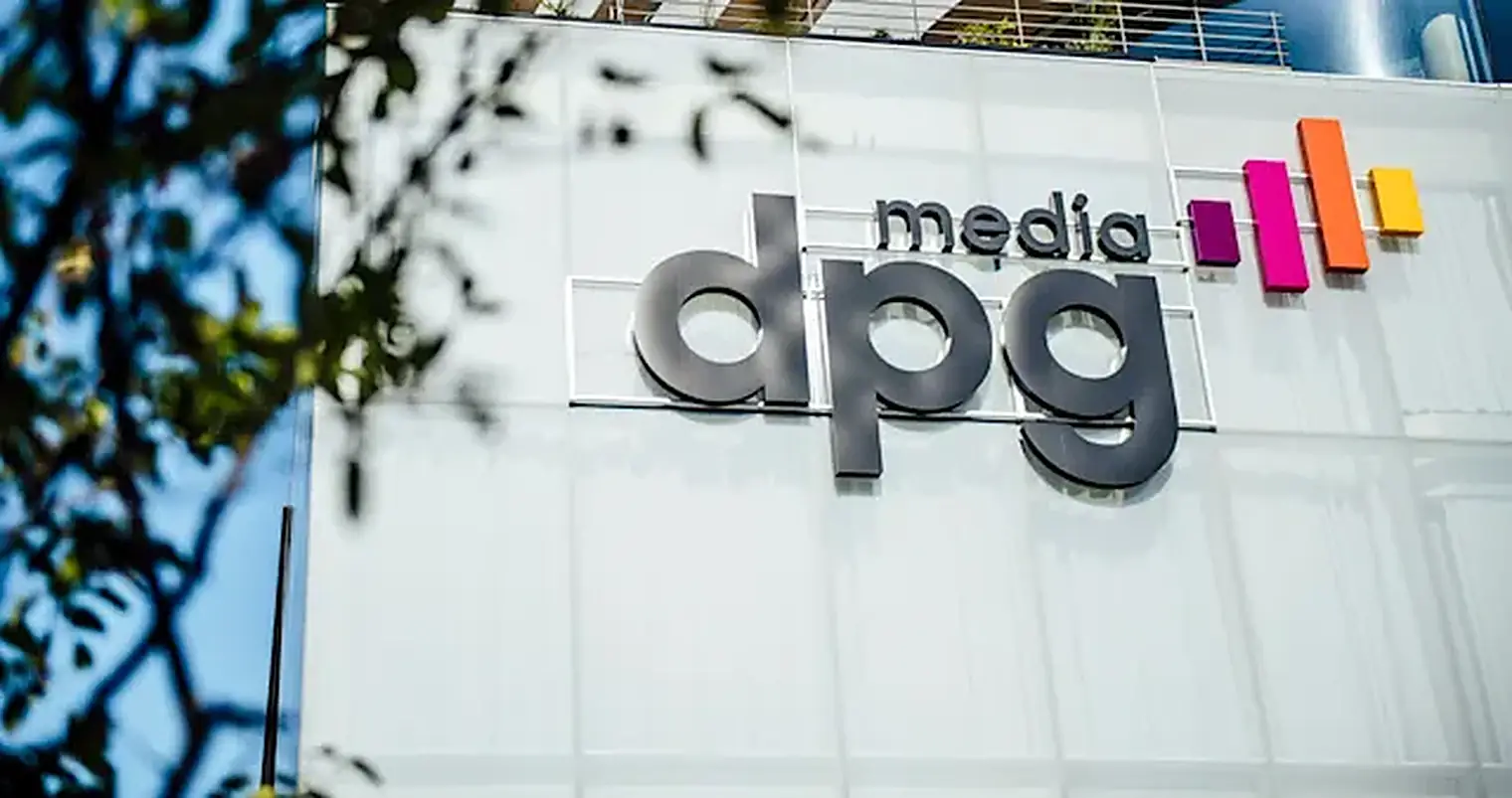 Het logo van dpg media in grote letters op een witte gevel, met bladeren van een boom op de voorgrond