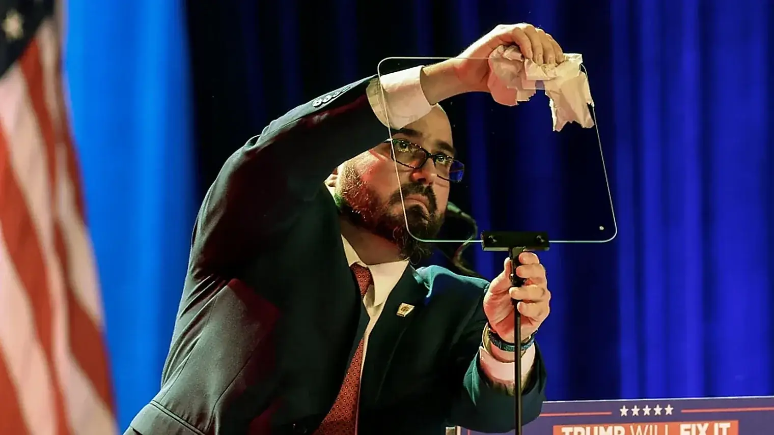 Een man in pak poetst met een doekje het glazen scherm van een teleprompter bij een spreekgestoelte, met een Amerikaanse vlag op de achtergrond