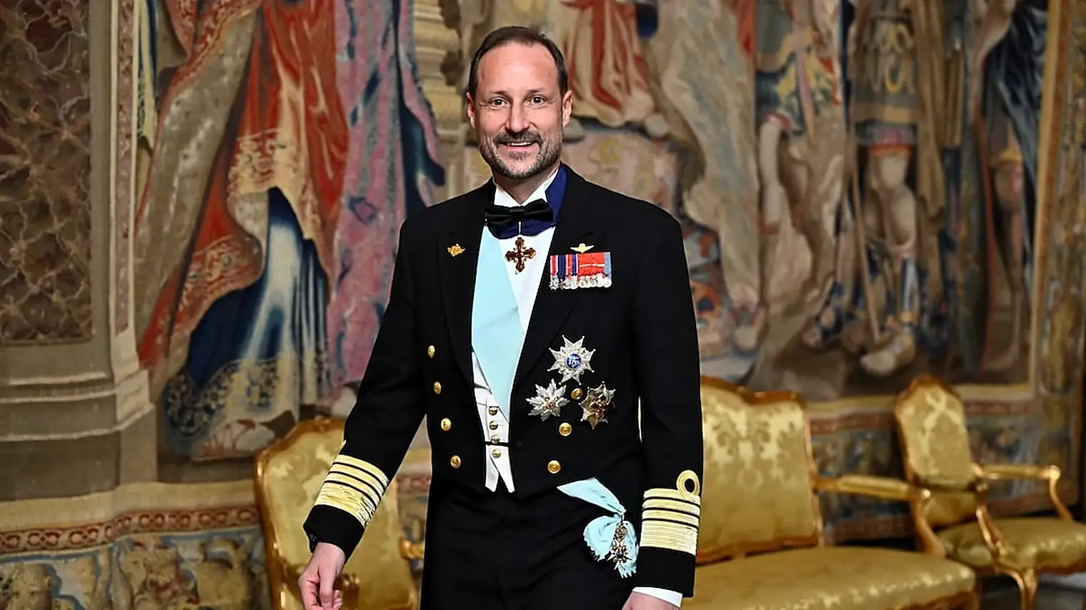 Haakon van Noorwegen in gala-uniform met onderscheidingen loopt door een zaal met wandtapijten en vergulde stoelen
