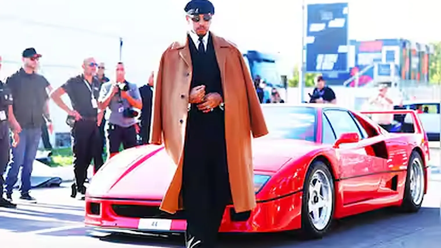 Hamilton loopt in pak en lange jas langs zijn rode Ferrari F40 terwijl fotografen en beveiligers bij de ingang van het paddock toekijken