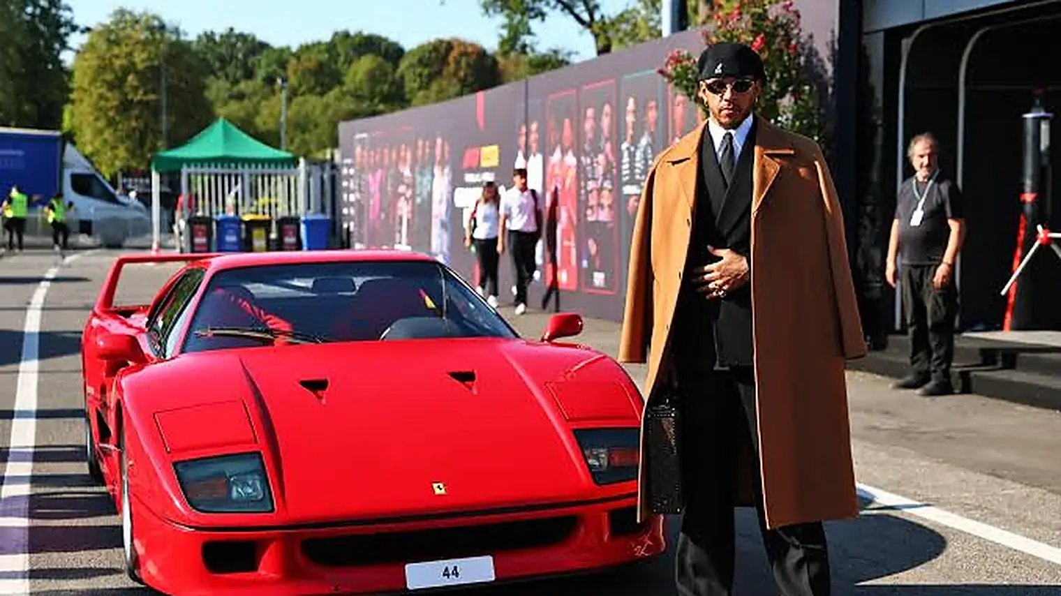Lewis Hamilton in een zwart pak met lange camelkleurige jas en pet naast een rode Ferrari F40 met het nummer 44 op de plaat, op het circuit van Monza