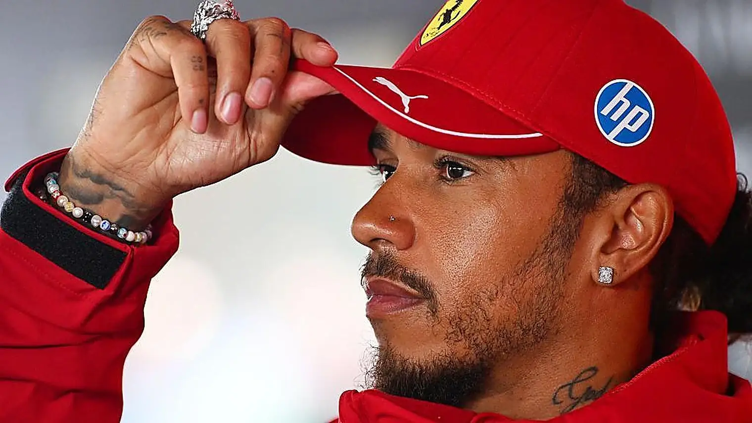 Lewis Hamilton in een rood Ferrari-jack tilt de rand van zijn rode pet op en kijkt langs de camera