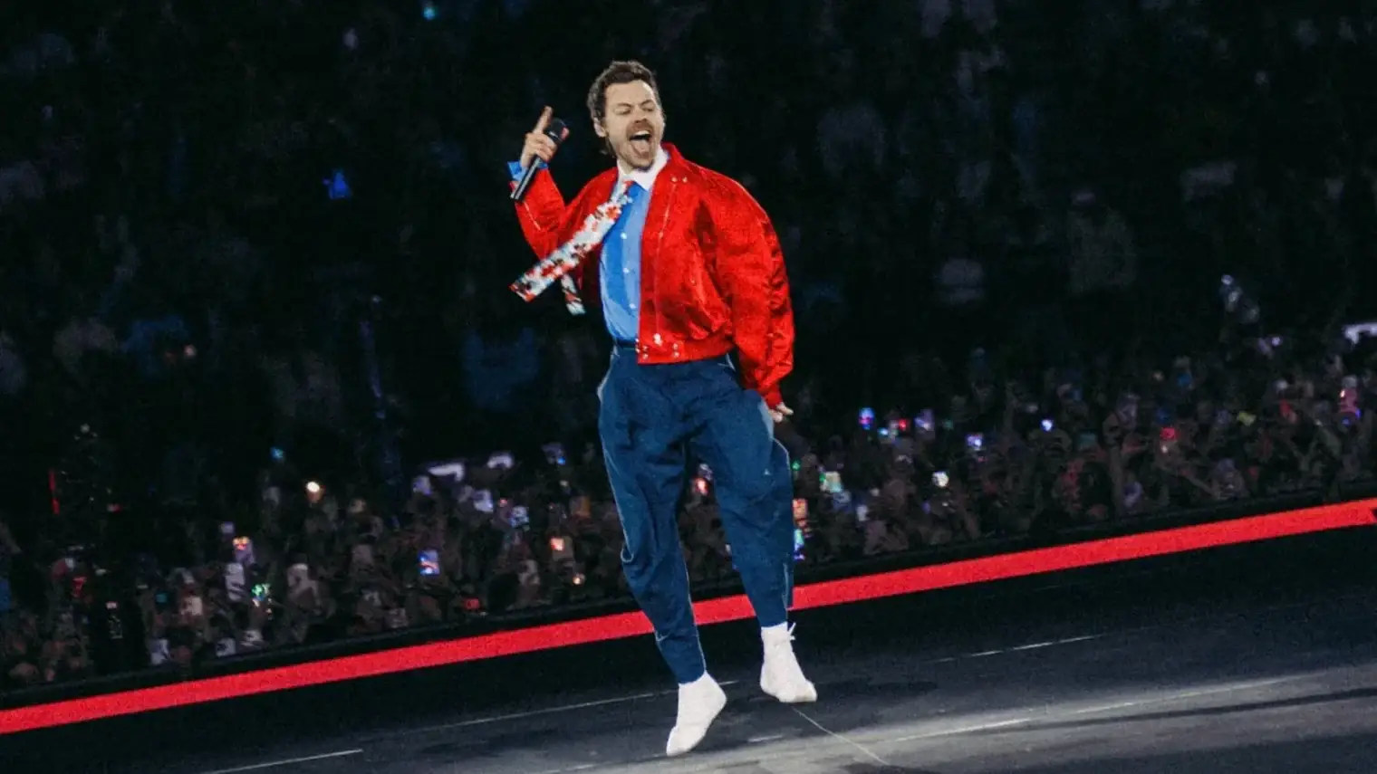 Harry Styles springt op het podium in een rode jas, met een microfoon in zijn hand en een uitzinnig publiek op de achtergrond