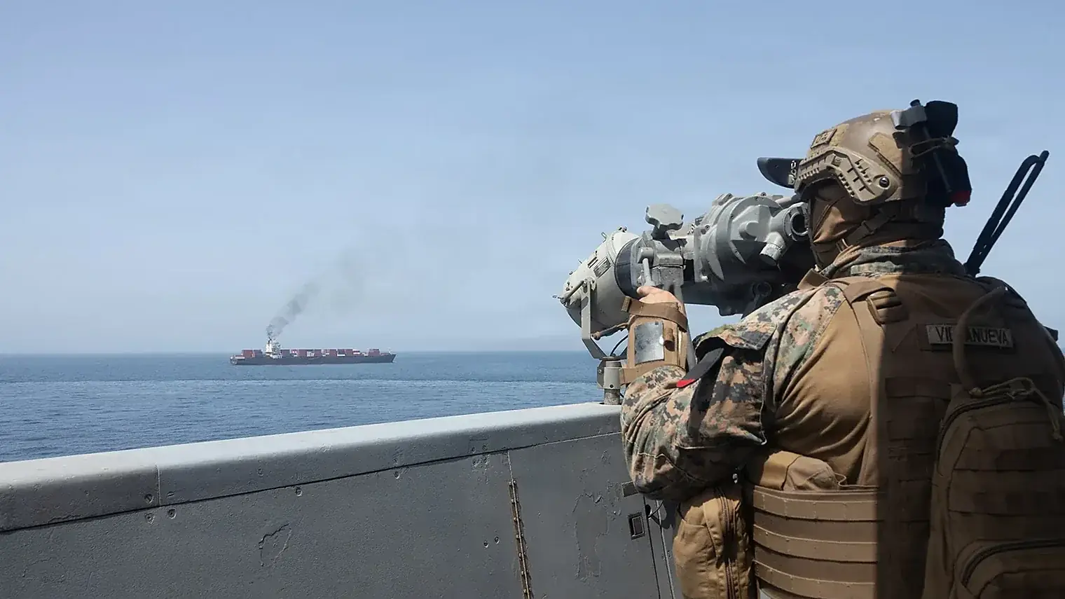 Een Amerikaanse marinier kijkt vanaf het dek van een oorlogsschip door een verrekijker naar een containerschip dat rook uitstoot op zee