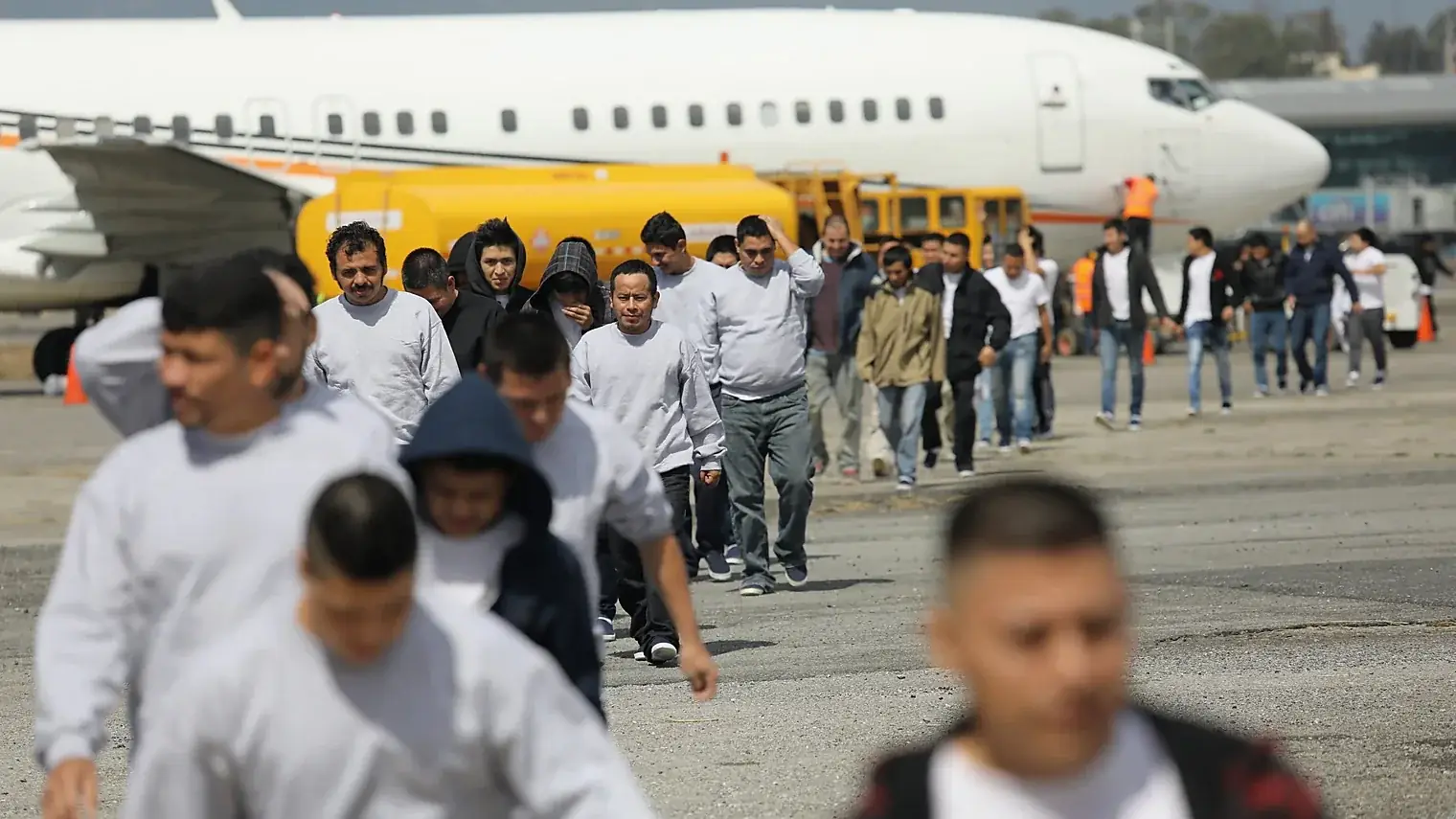 Een rij mannen in grijze truien loopt over een platform weg van een wit vliegtuig na een deportatievlucht