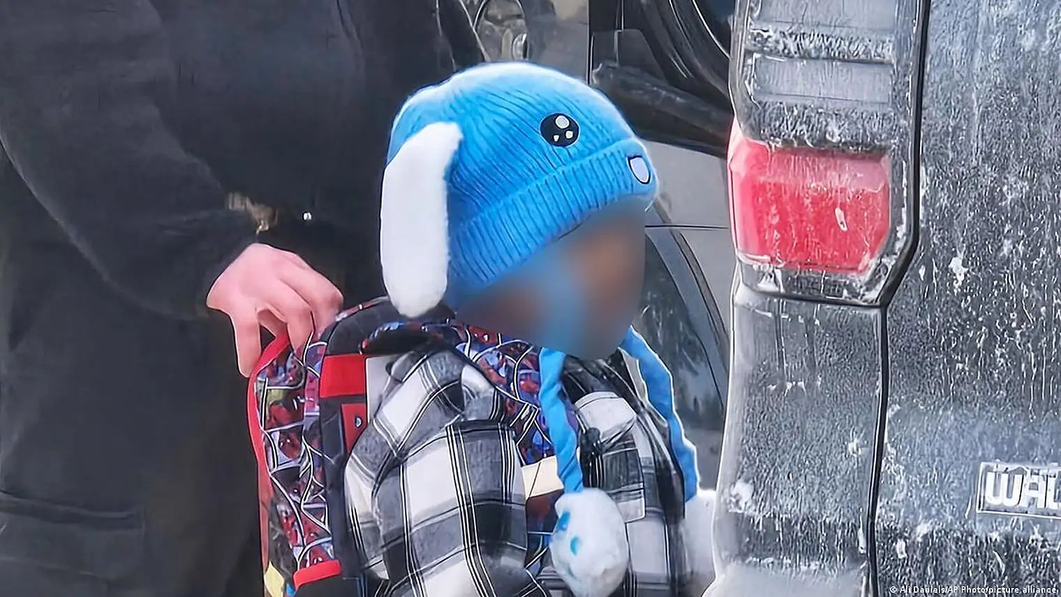 Een klein kind met een blauwe muts en een rugzak met superheldenprint wordt bij een auto meegenomen, het gezicht is onherkenbaar gemaakt