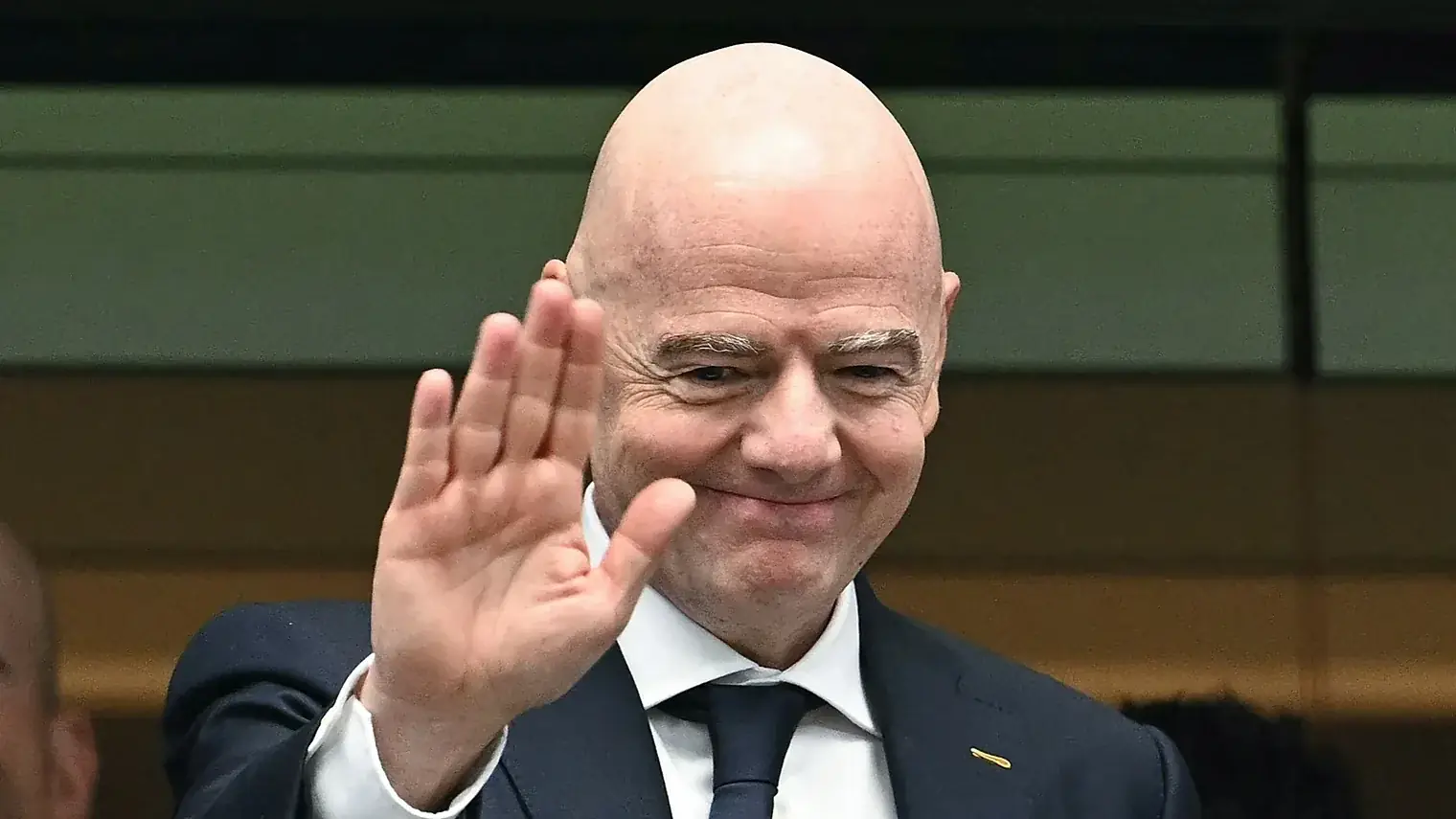 FIFA-voorzitter Gianni Infantino in donker pak zwaait met zijn hand, met op de achtergrond een tribune