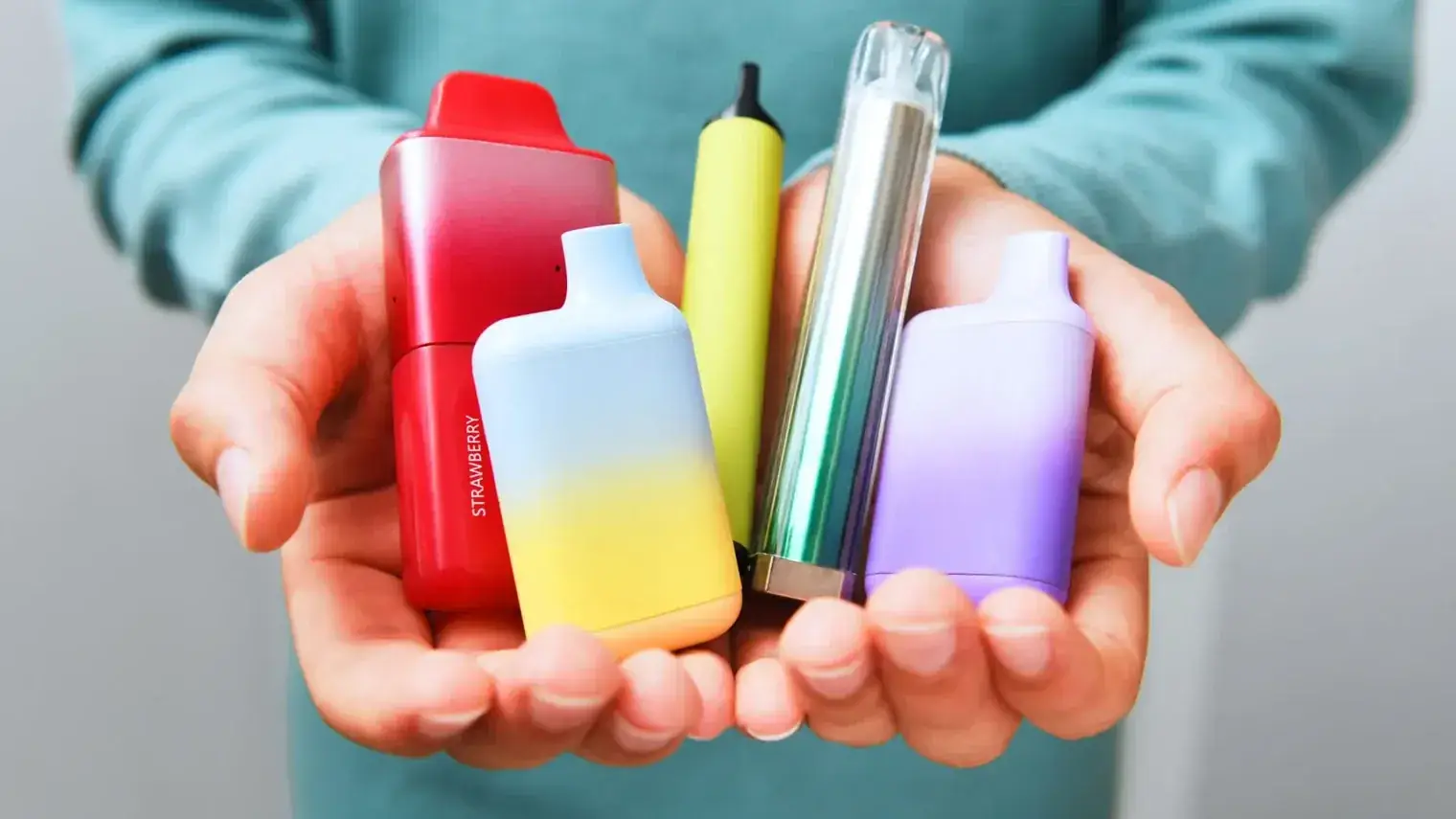 Twee handen houden vijf kleurrijke vapes vast voor een groenblauwe trui