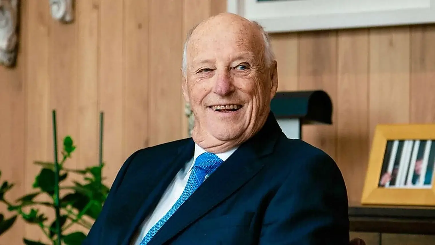 Koning Harald van Noorwegen lacht in een donkerblauw pak met lichtblauwe stropdas, zittend voor een houten wand