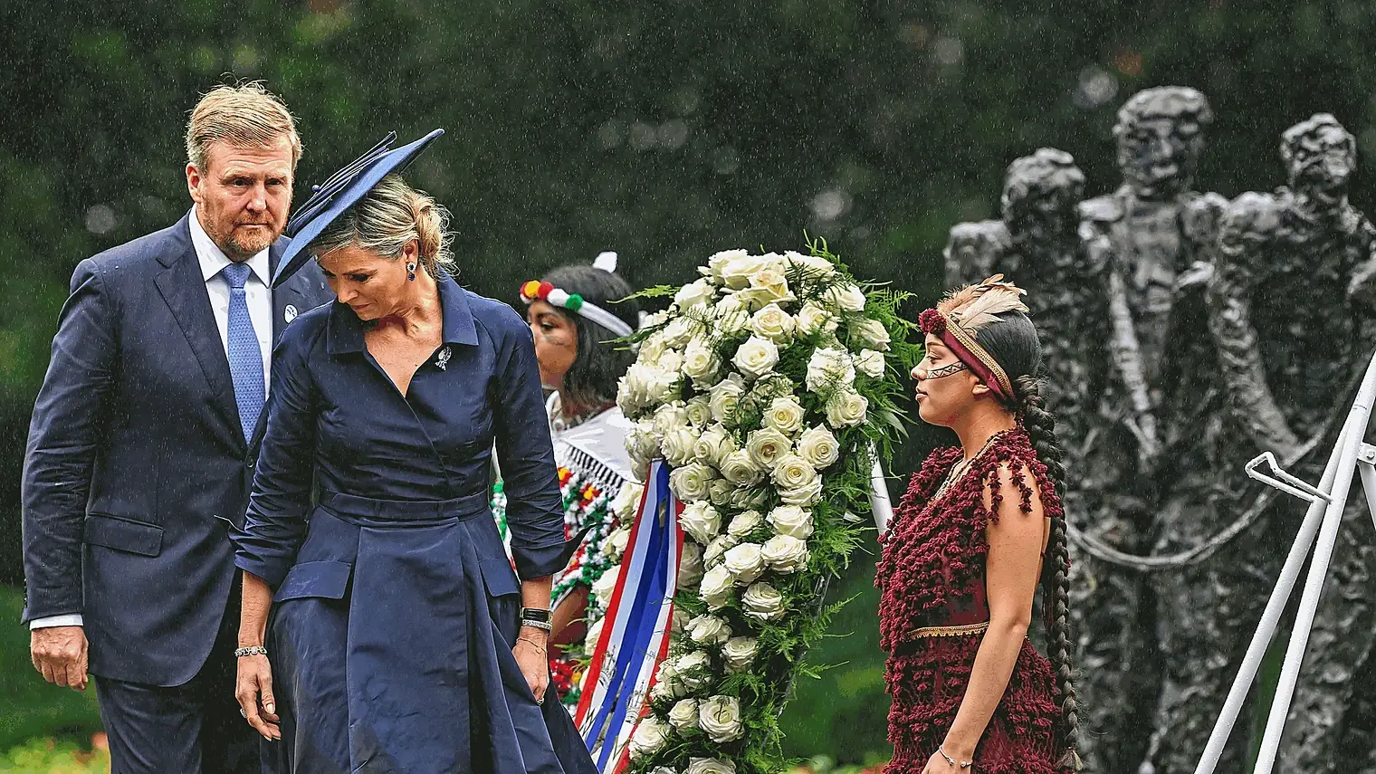 Koning Willem-Alexander en koningin Maxima in de regen naast een krans van witte rozen