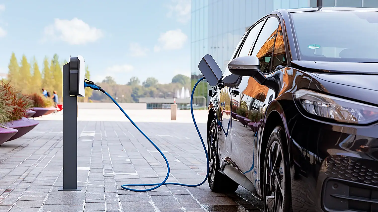 Elektrische auto met een blauwe laadkabel aan een laadzuil op een parkeerplaats bij een kantoorgebouw van glas