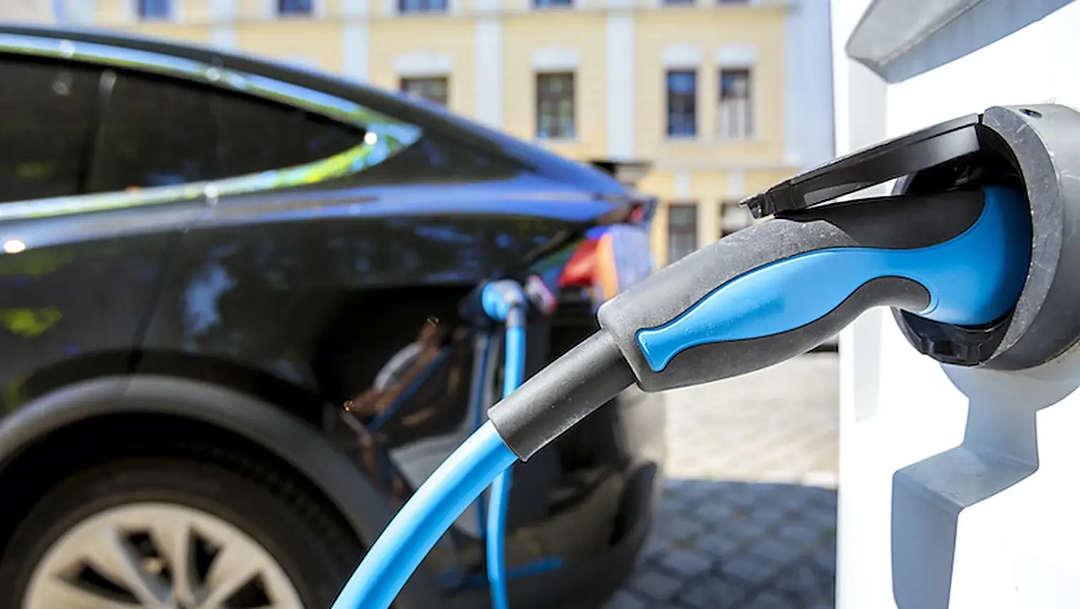 Blauwe laadkabel met stekker in een openbare laadpaal, met een zwarte elektrische auto en een woonblok op de achtergrond