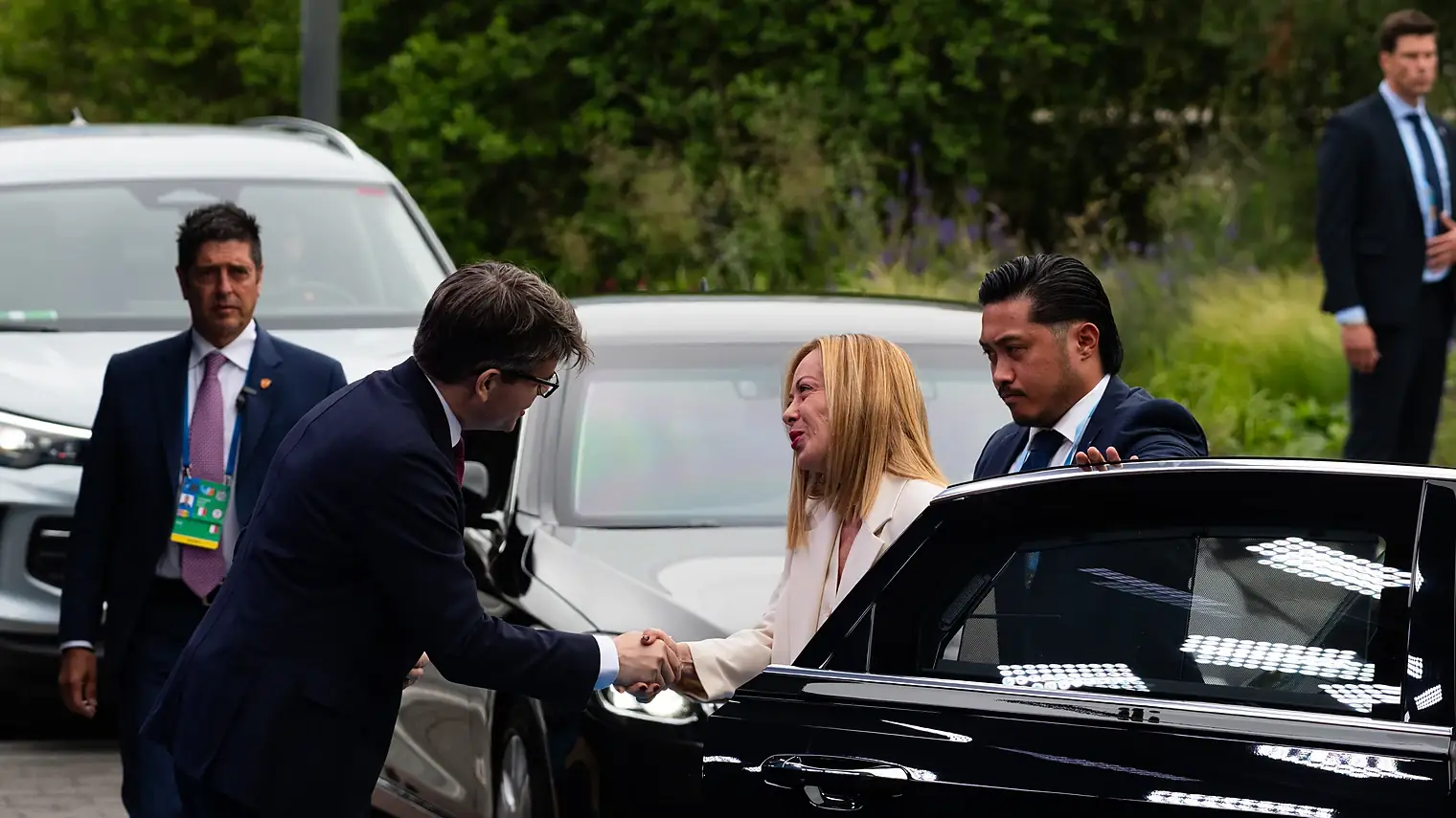 Giorgia Meloni in een crèmekleurig pak schudt een hand bij haar aankomst met de auto op een NAVO-top, met beveiligers eromheen