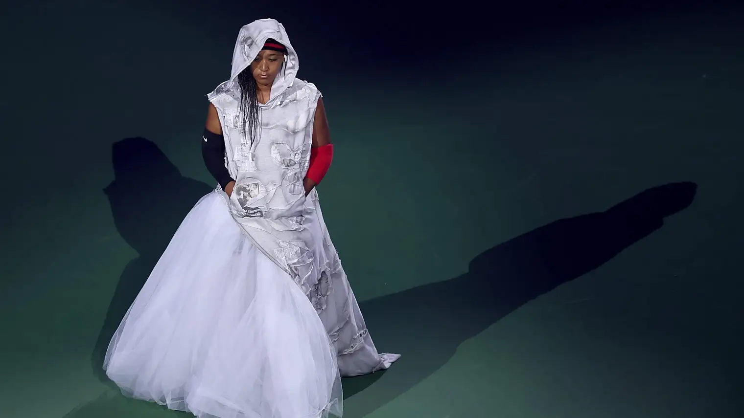 Naomi Osaka loopt in een witte jurk met capuchon en lange tulerok over de groene baan van het Arthur Ashe Stadium