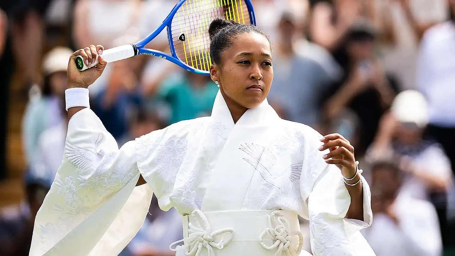 Osaka met haar racket over haar schouder in een witte kimonojas met geborduurde kraanvogels en bloesems en een geknoopte obi-band