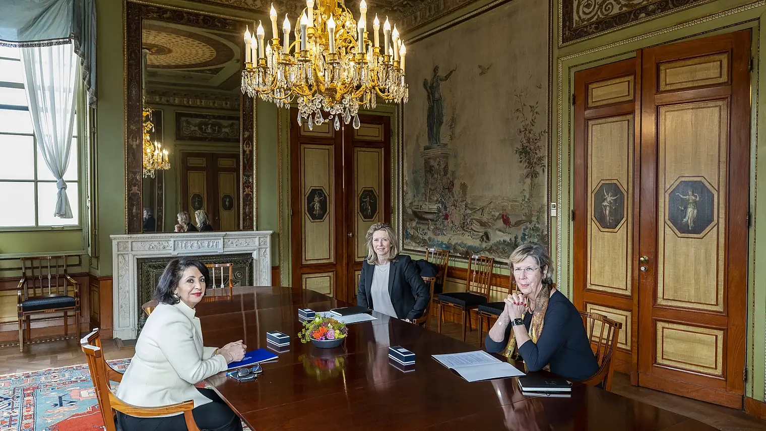 Drie vrouwen op ruime afstand van elkaar aan een lange vergadertafel in een zaal met kroonluchter en wandtapijt