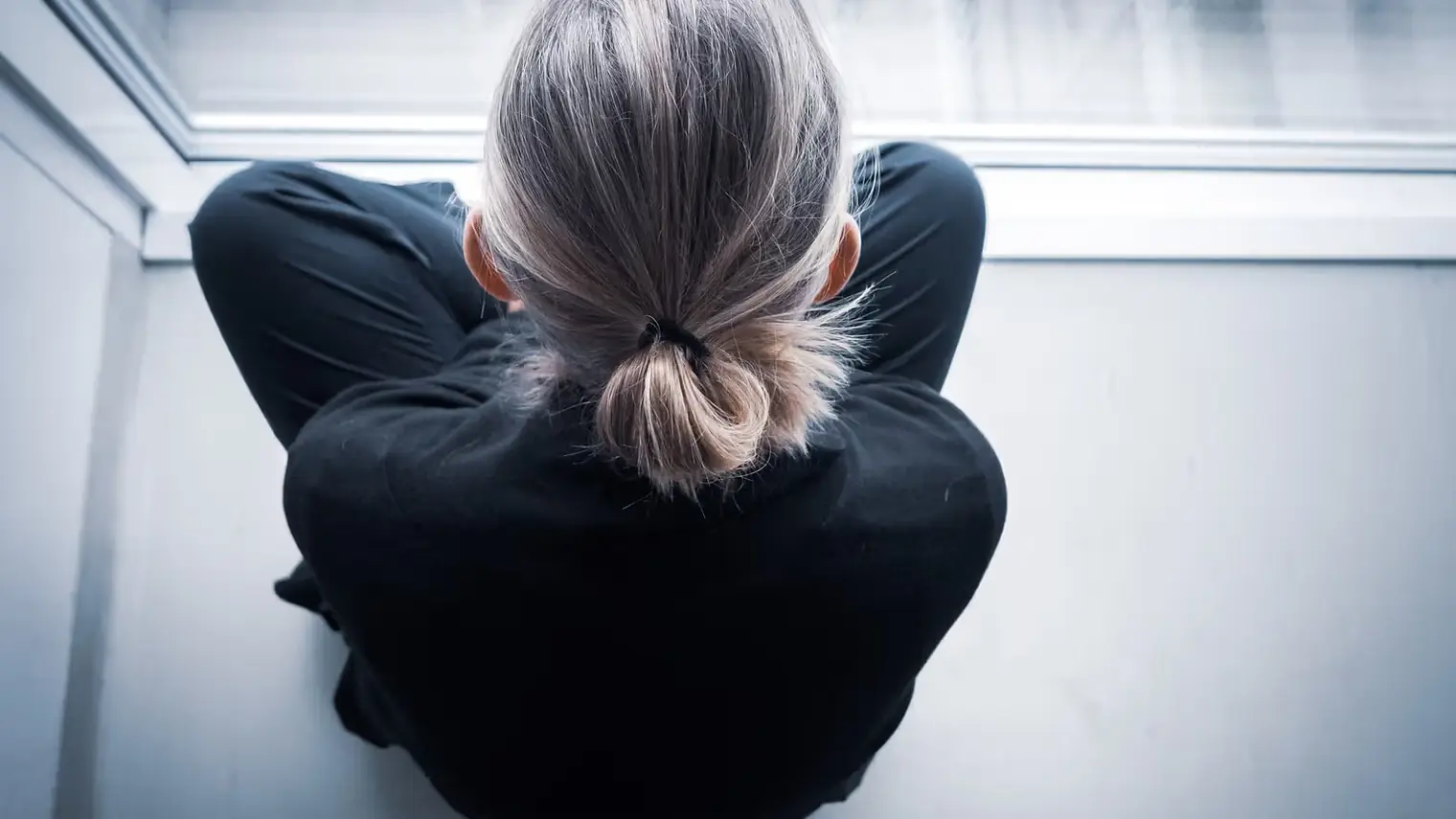 Een persoon zit met opgetrokken knieën bij een raam, het gezicht niet zichtbaar