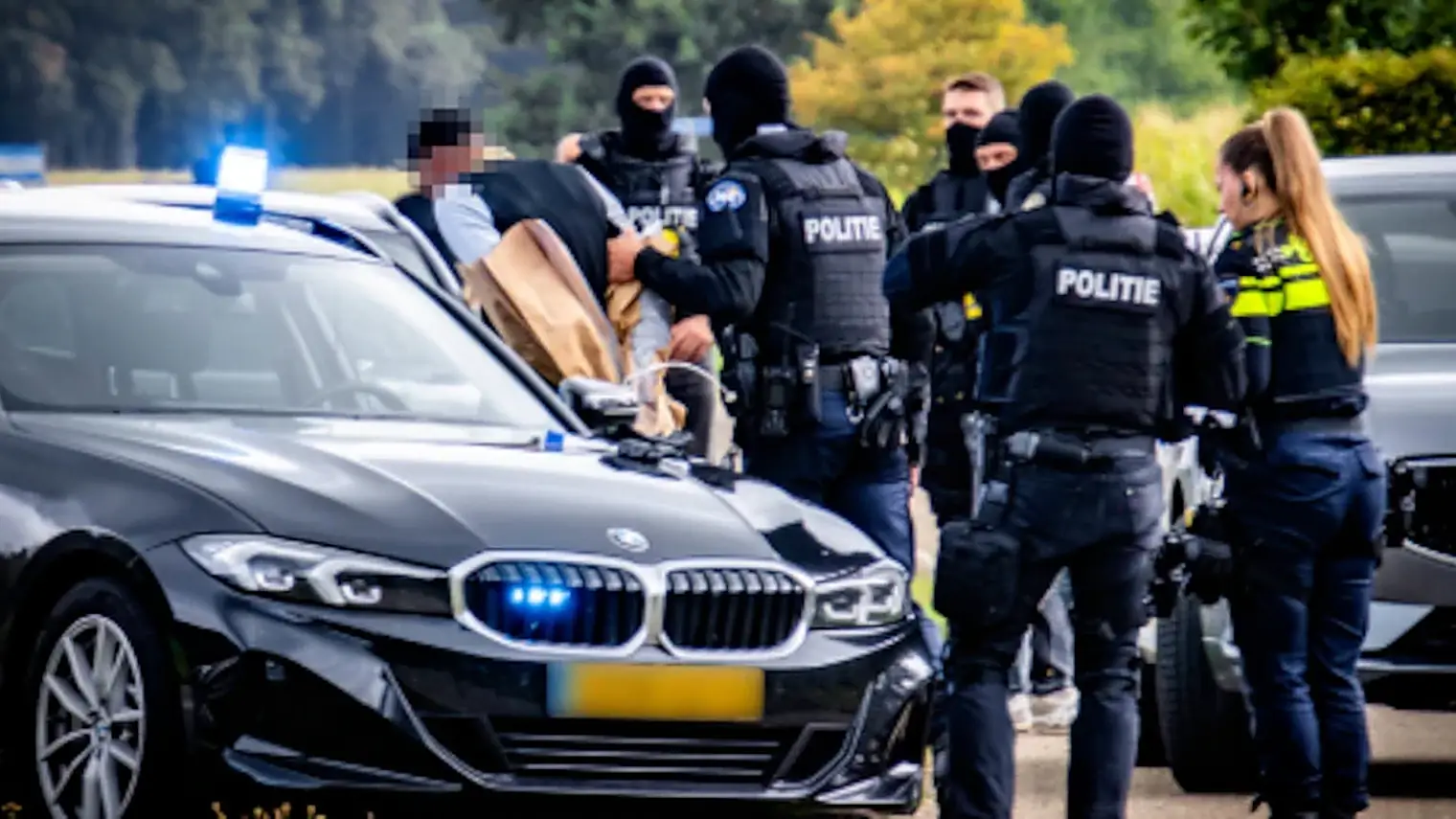 Agenten in kogelwerende vesten en bivakmutsen begeleiden een aangehouden persoon langs een onopvallende politieauto met blauw zwaailicht