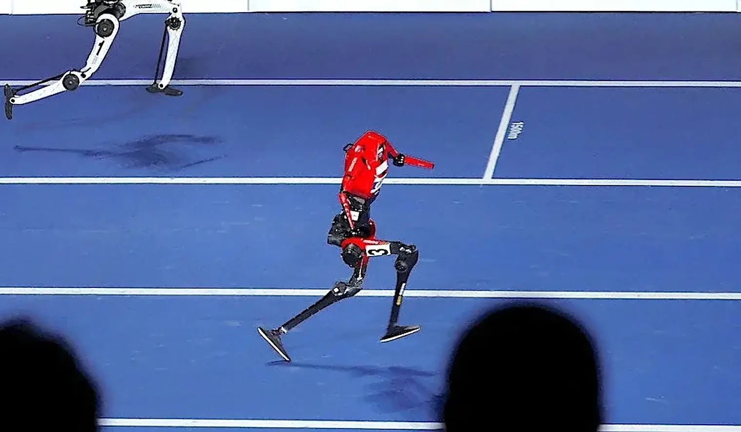 Rode humanoide robot sprint over een blauwe atletiekbaan in een hal, met een witte robot een baan verder achter zich