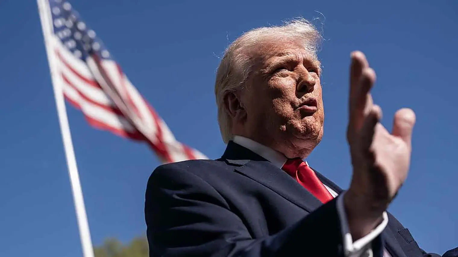 President Trump gebaart tijdens het spreken, van onderop gefotografeerd met de Amerikaanse vlag en een blauwe lucht achter hem