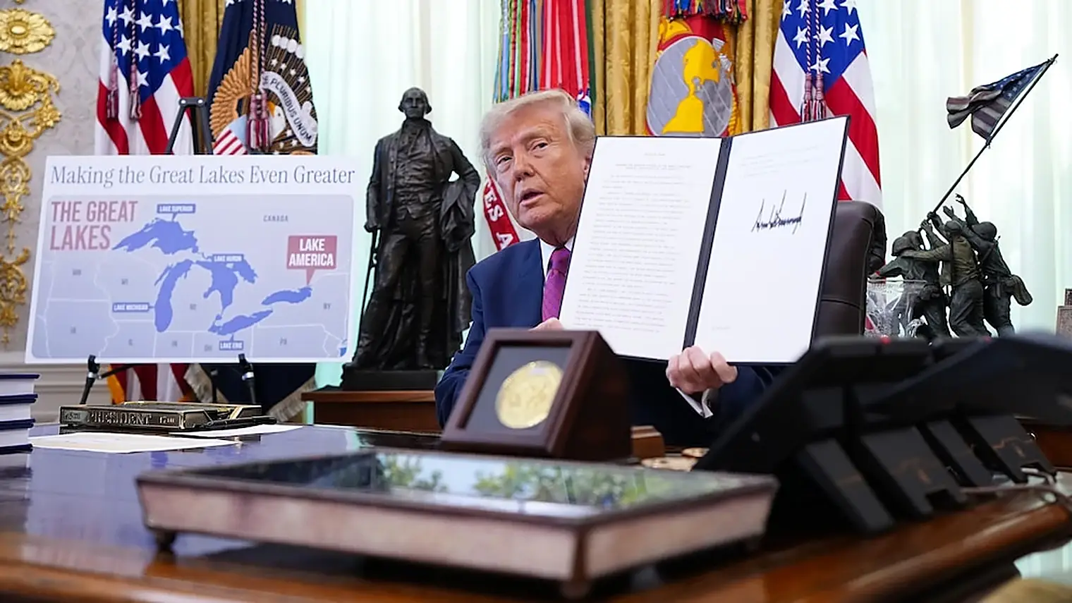 Trump houdt in het Oval Office het ondertekende decreet omhoog, naast een bord met een kaart van de Grote Meren en het opschrift Lake America