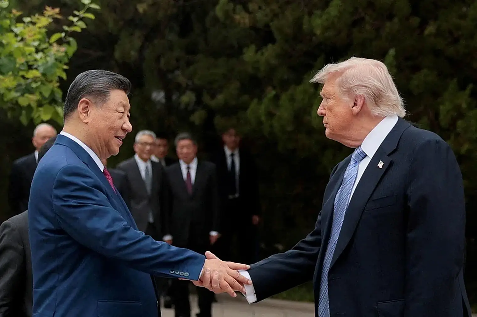 Xi Jinping en Donald Trump geven elkaar buiten een hand, met een delegatie in donkere pakken op de achtergrond tussen het groen